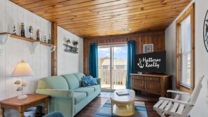 A cozy living room with a wooden ceiling, a green sofa, a small table with a lamp, a television on a wooden cabinet, and a view through a sliding glass door.