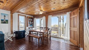 A wooden-floored room with a dining table, chairs, a bookshelf, and a view through large windows.