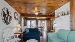A wooden interior with a ceiling fan, a blue sofa, a white chair, and a dining area.