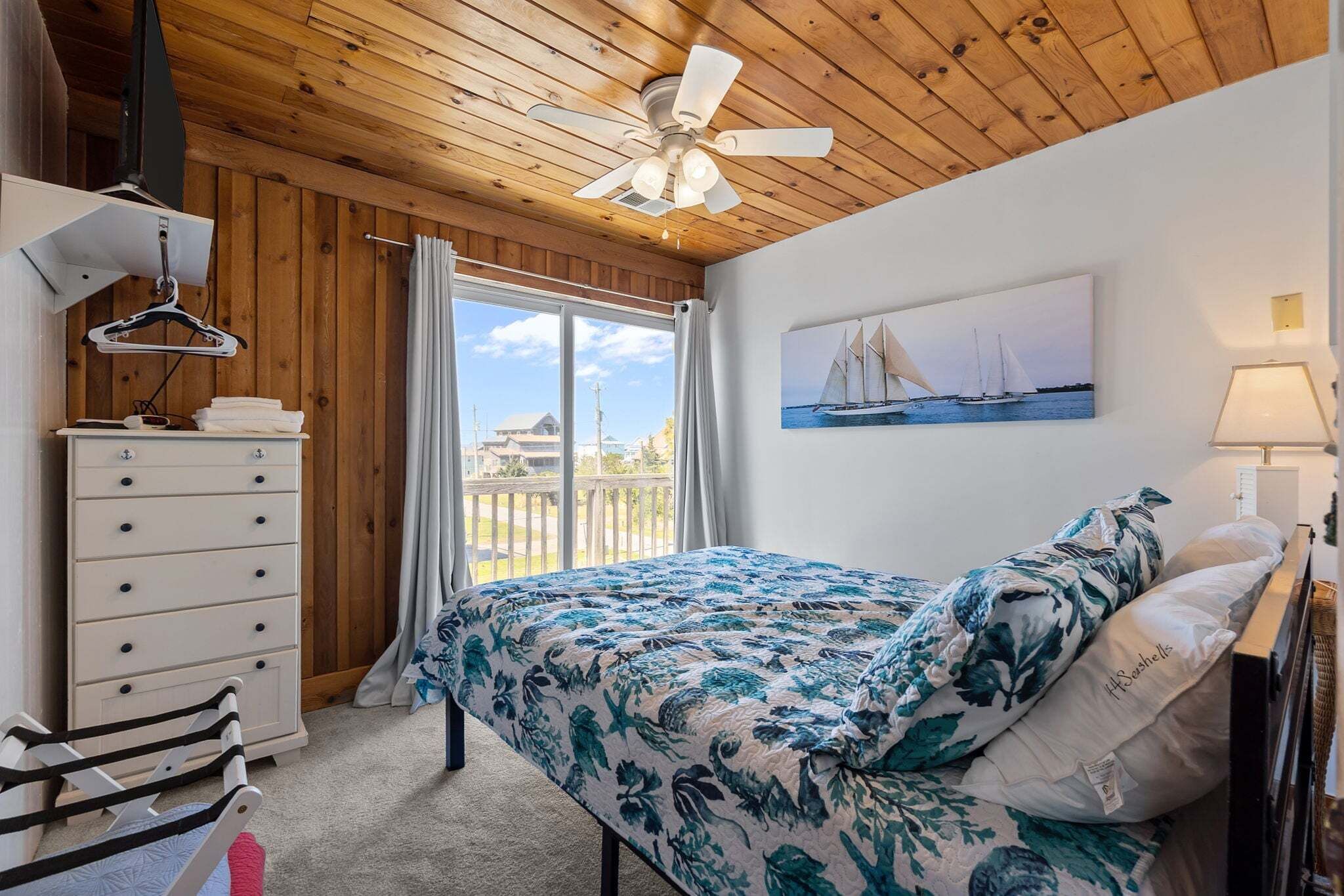 A bedroom with a bed, a dresser, a hanging clothes rack, a ceiling fan, a window with curtains, and a painting of sailboats.