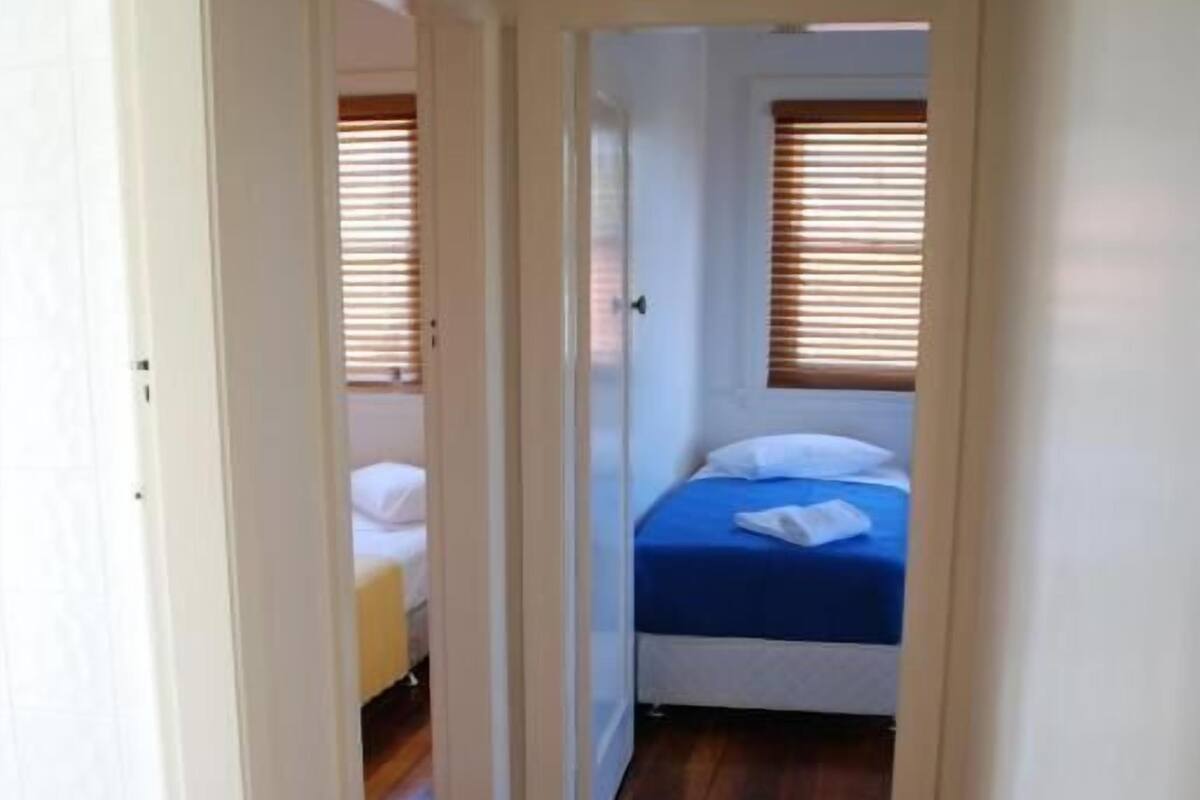A narrow hallway with wooden flooring leading to a bedroom with a bed and a window with blinds.