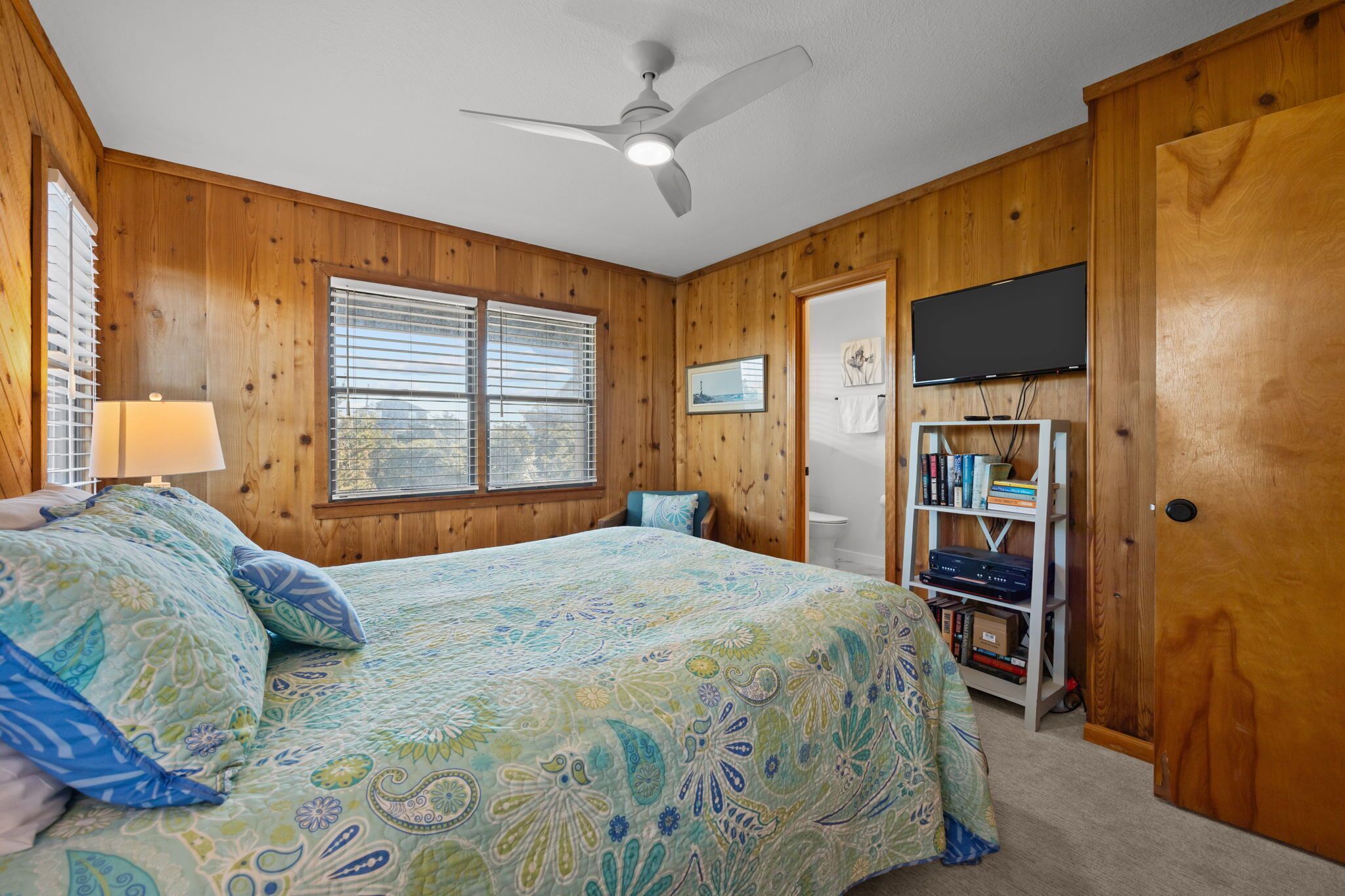A bedroom with a bed, a ceiling fan, a TV mounted on the wall, and a bookshelf.