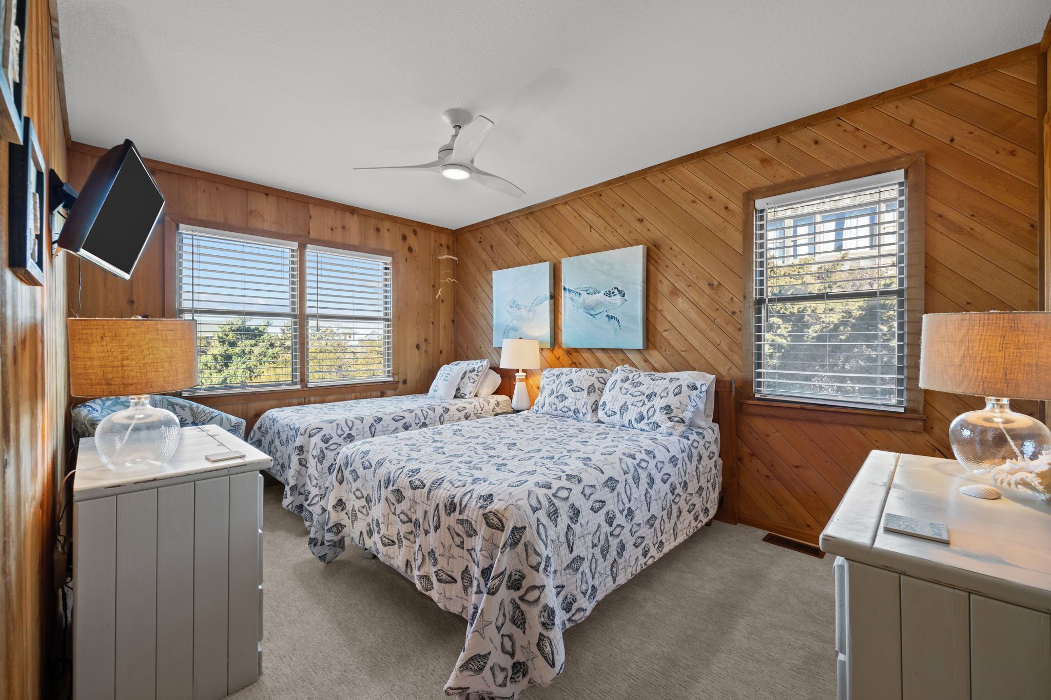 A bedroom with two beds, wooden walls, a ceiling fan, and two windows with blinds.