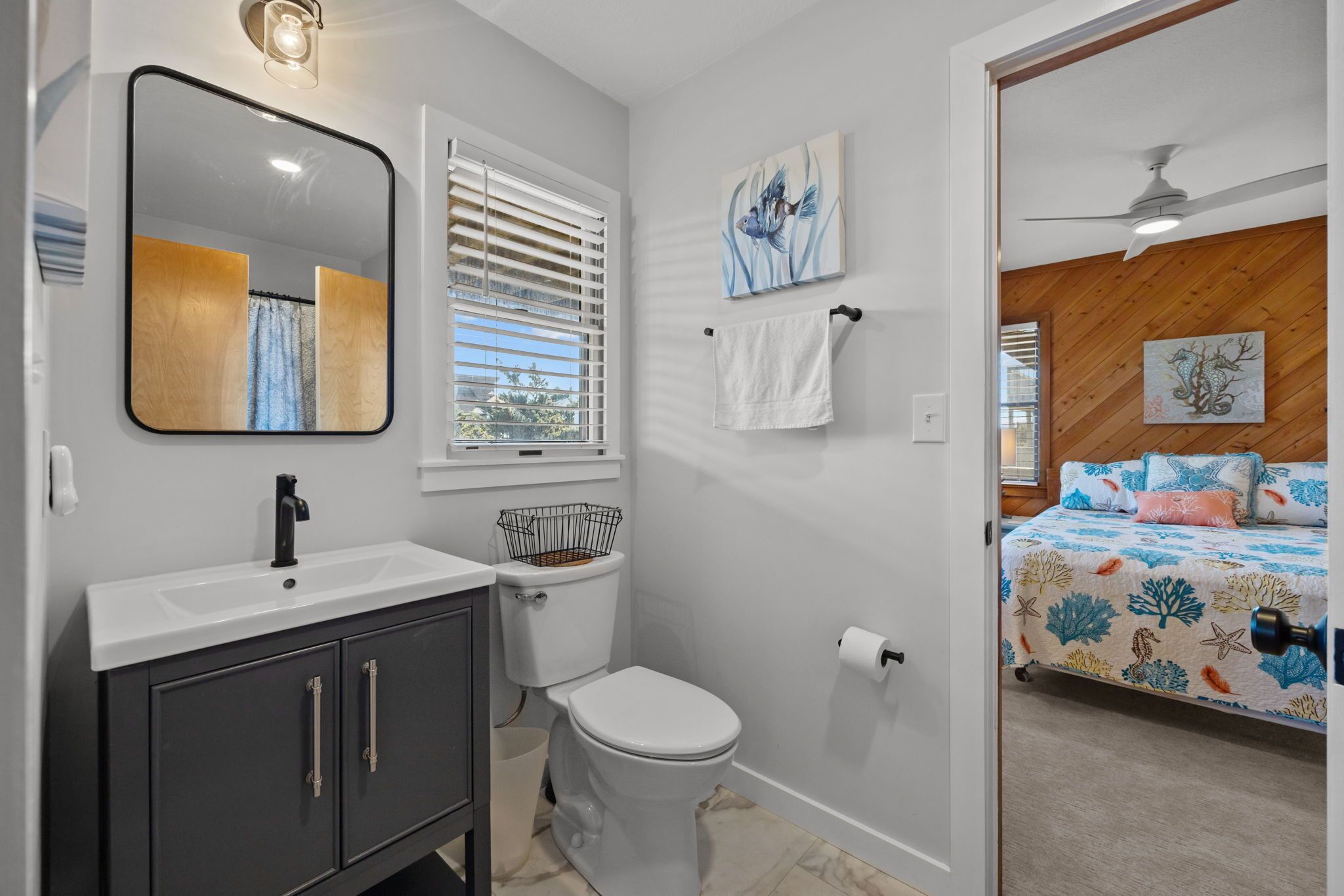 A bathroom with a white sink, black cabinet, and a toilet. There is a window with blinds, a towel rack, and a painting on the wall.