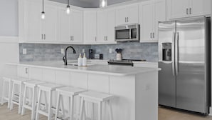 A modern kitchen with white cabinets, a stainless steel refrigerator, a microwave, and a bar area with stools. - Stunning sound view home, close to the beach in the heart of Surf City (Surf City)