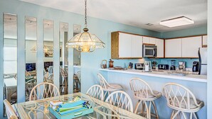 A dining area with wicker chairs and a glass table, a kitchen with white cabinets and appliances, and a hanging light fixture. - Ocean and waterway-view duplex - pool, beach access, washer/dryer, central AC (Topsail Beach)