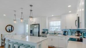 A modern kitchen with white cabinets, a large island with bar stools, a refrigerator, a microwave, and a coffee maker. - Oceanfront home with beach access, a game room, washer/dryer, & central AC (Topsail Beach)