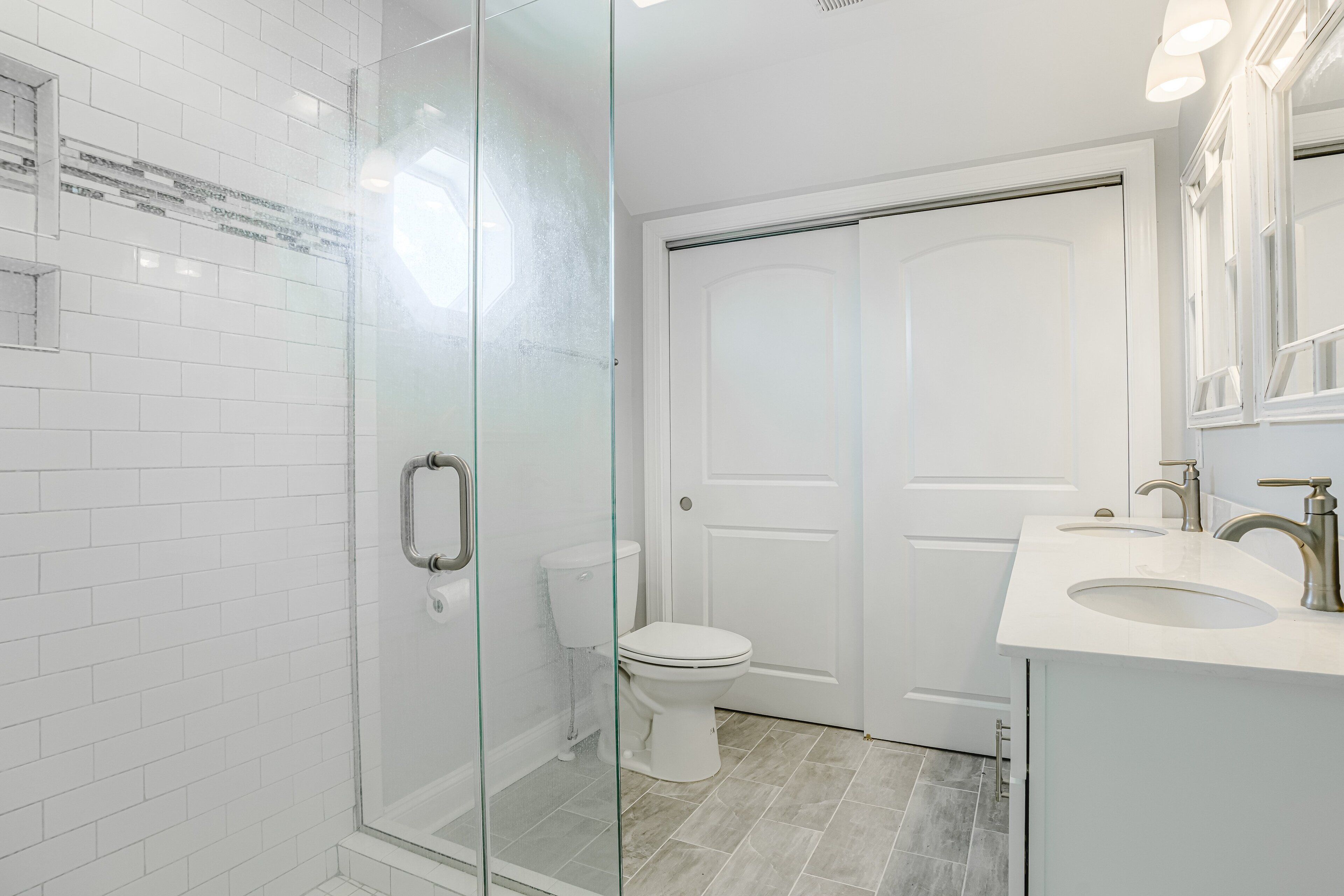A modern bathroom with white tiled walls, a glass shower enclosure, a white toilet, and a vanity with a sink and mirror.