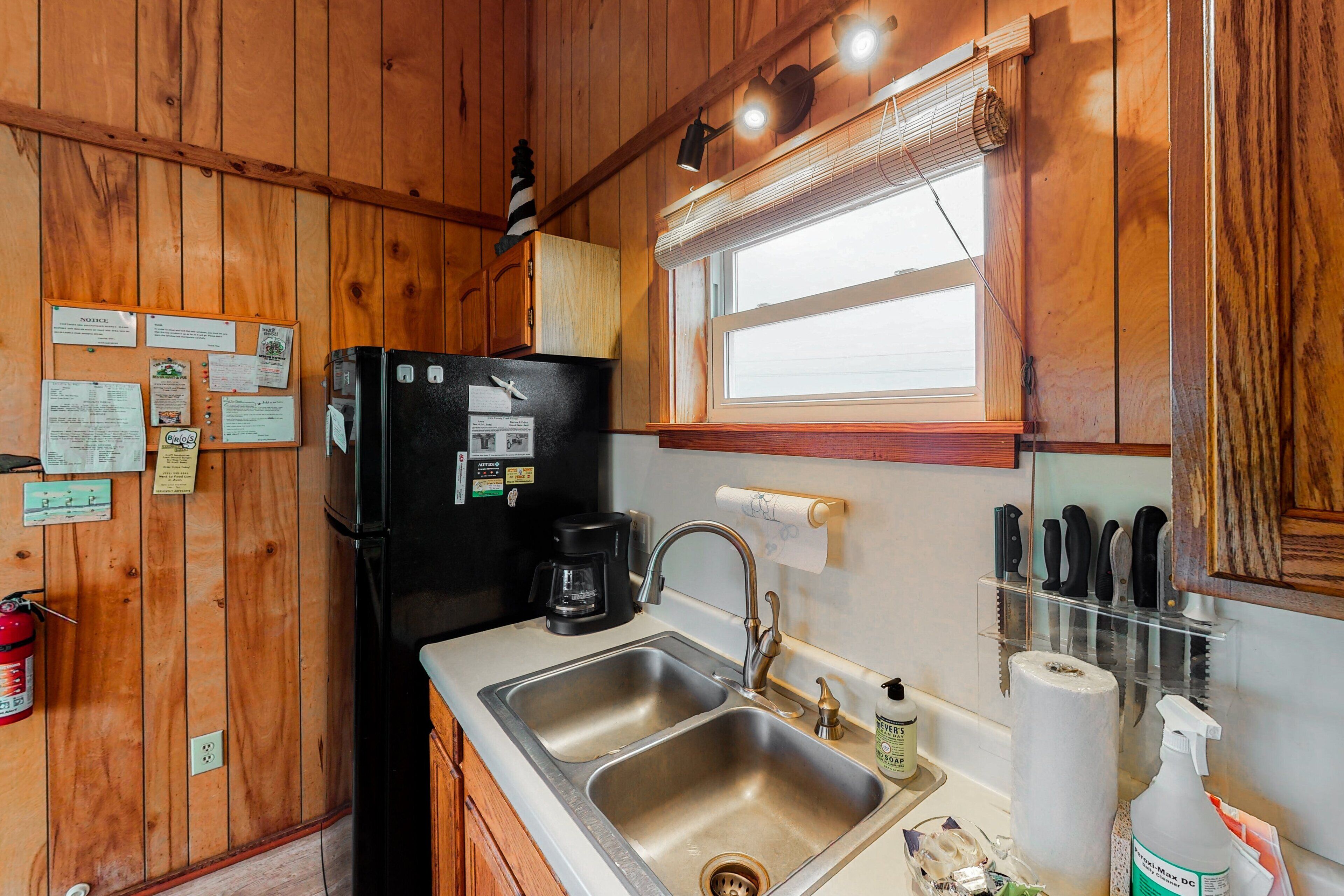 A compact kitchen with wooden walls, a black refrigerator, a coffee maker, a sink, and a window with blinds.