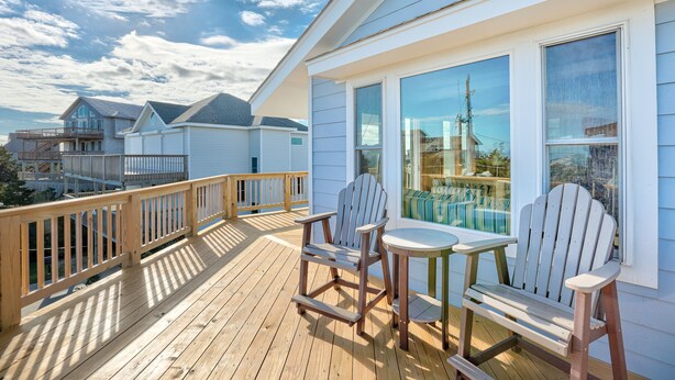 Eine Holzterrasse mit zwei Adirondack-Stühlen und einem kleinen Tisch, mit Blick auf einen Pool und benachbarte Häuser.