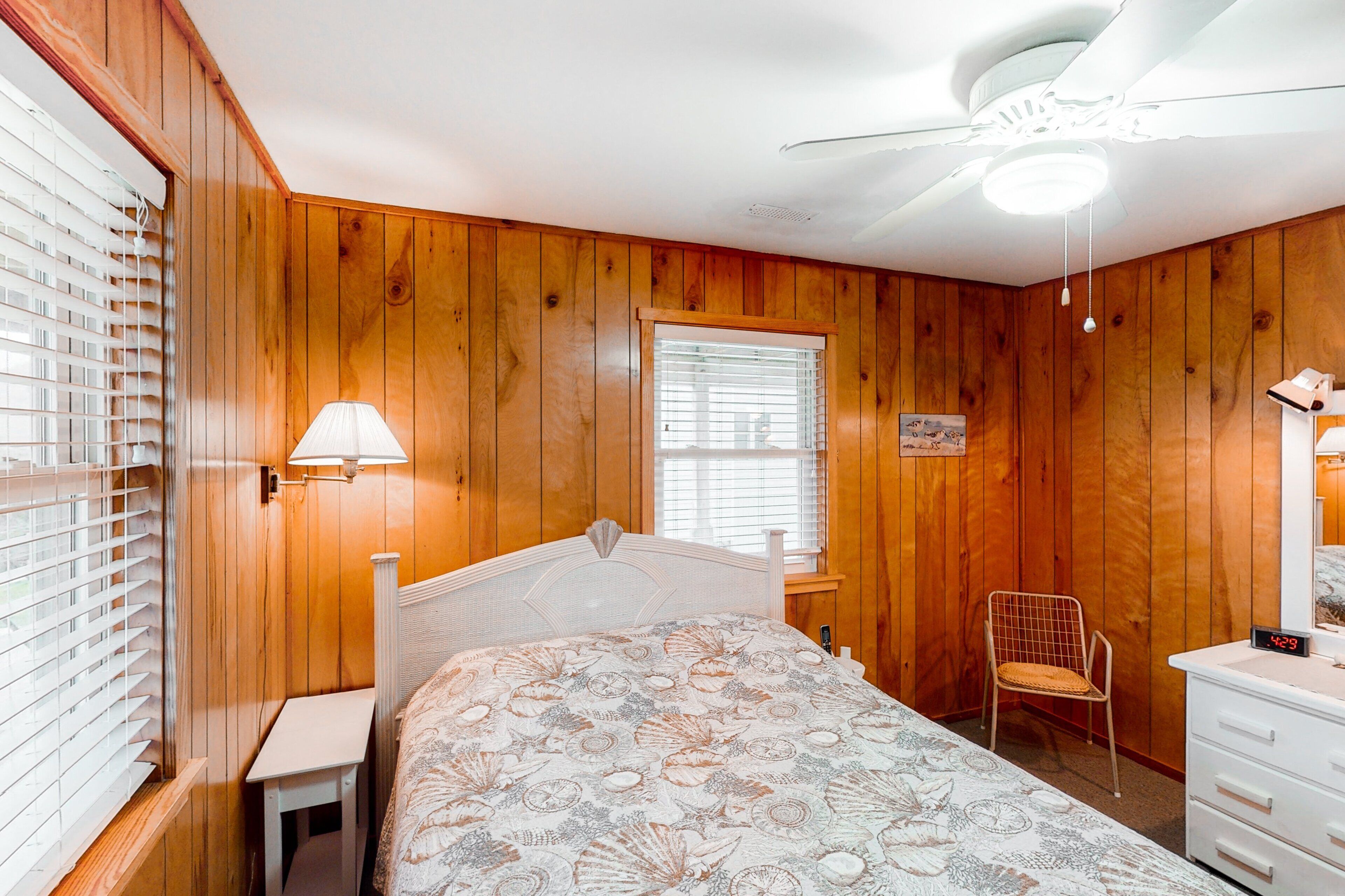 A bedroom with wooden walls, a bed with a floral bedspread, a bedside table, a chair, a dresser, a window with blinds, and a ceiling fan.