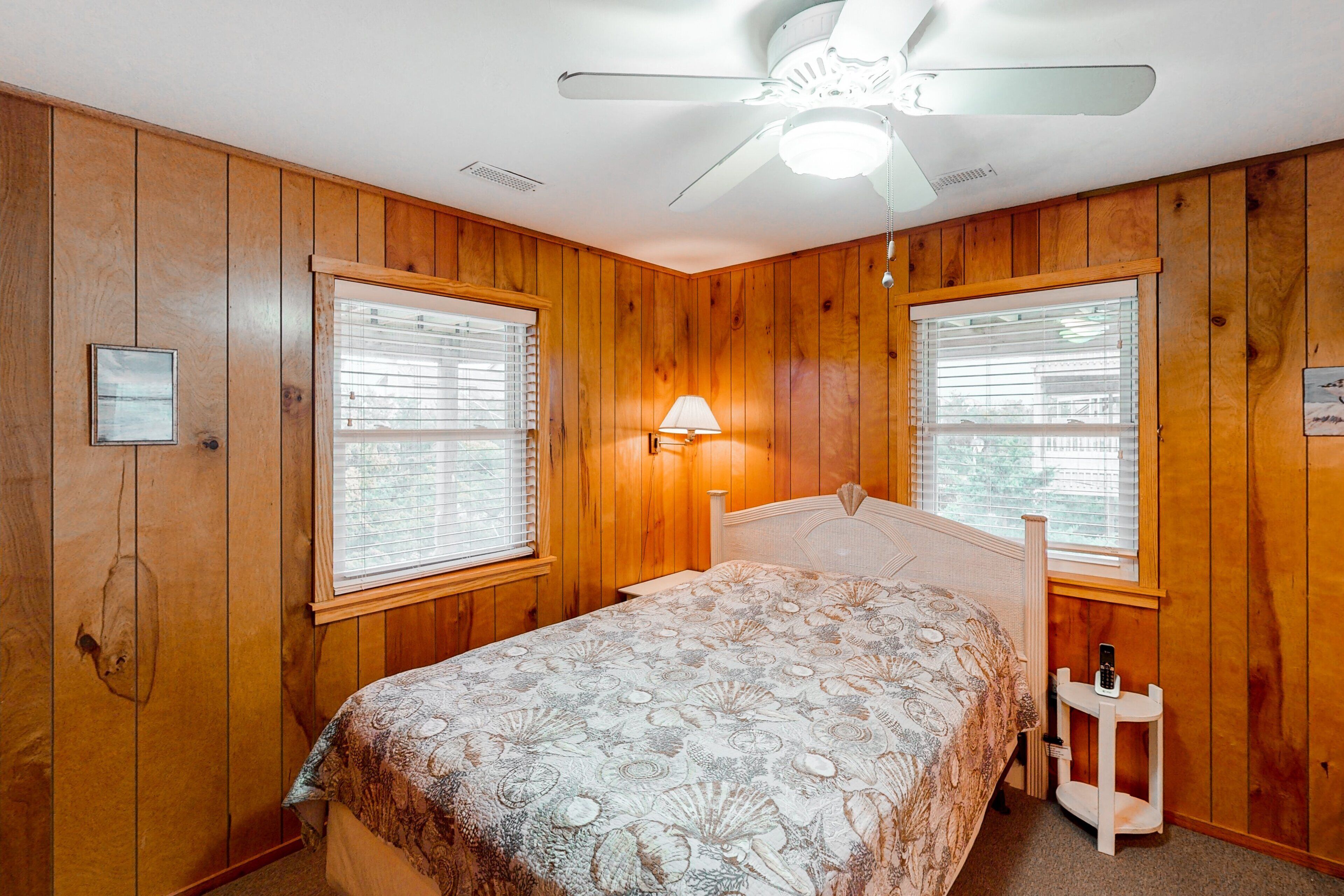 A bedroom with wooden paneling, a bed with a patterned bedspread, two windows with blinds, a ceiling fan, and a nightstand with a lamp.