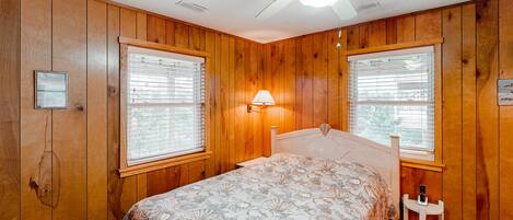 A bedroom with wooden paneling, a bed with a patterned bedspread, two windows with blinds, a ceiling fan, and a nightstand with a lamp.