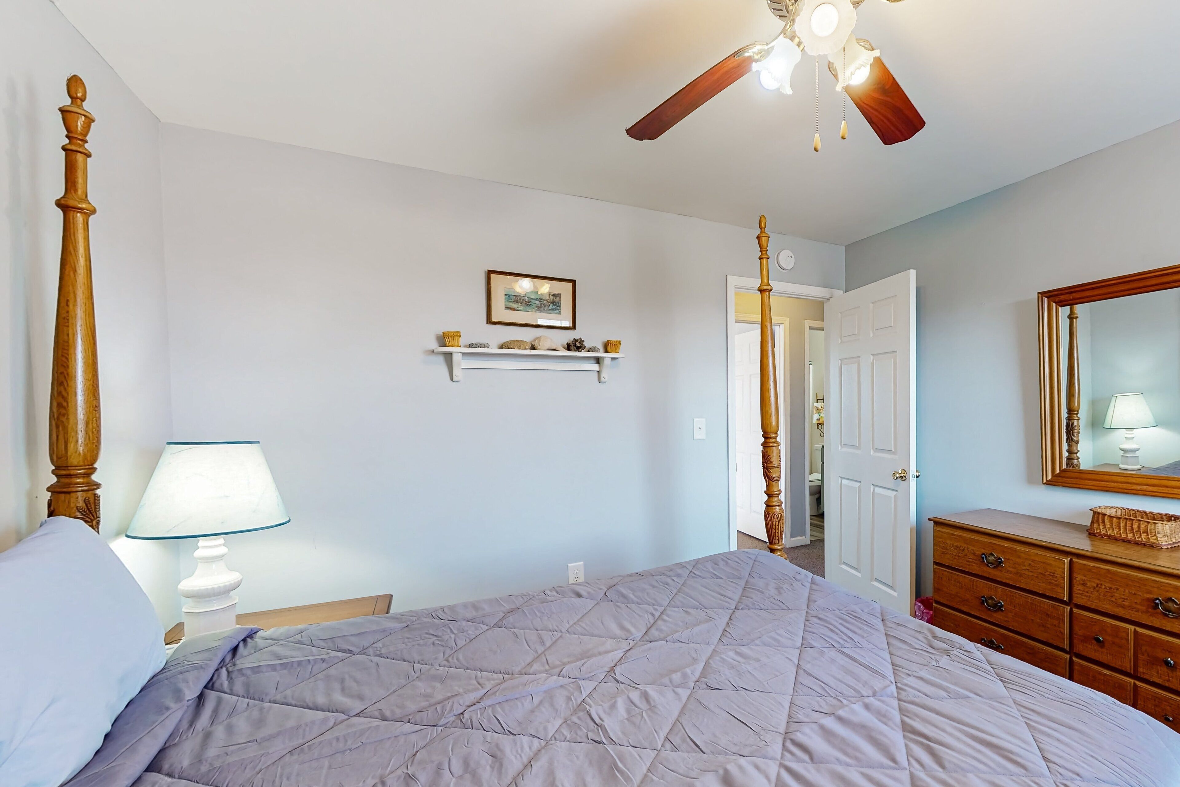 A bedroom with a bed, a nightstand, a lamp, a dresser, a mirror, a ceiling fan, and a door leading to another room.