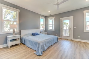 Un dormitorio con cama, mesitas de noche, ventilador de techo y ventanas amplias.