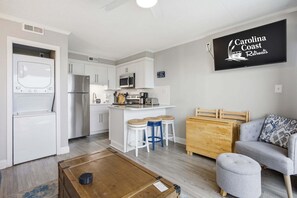 A modern kitchen with white cabinets, a microwave, and a refrigerator. There is a small island with stools, a wooden chest of drawers, and a grey sofa with patterned cushions. A flat-screen TV is mounted on the wall displaying 'Carolina Coast Retreats'. - Oceanfront condo with great view, fast WiFi, & central AC - steps to the beach (Carolina Beach)