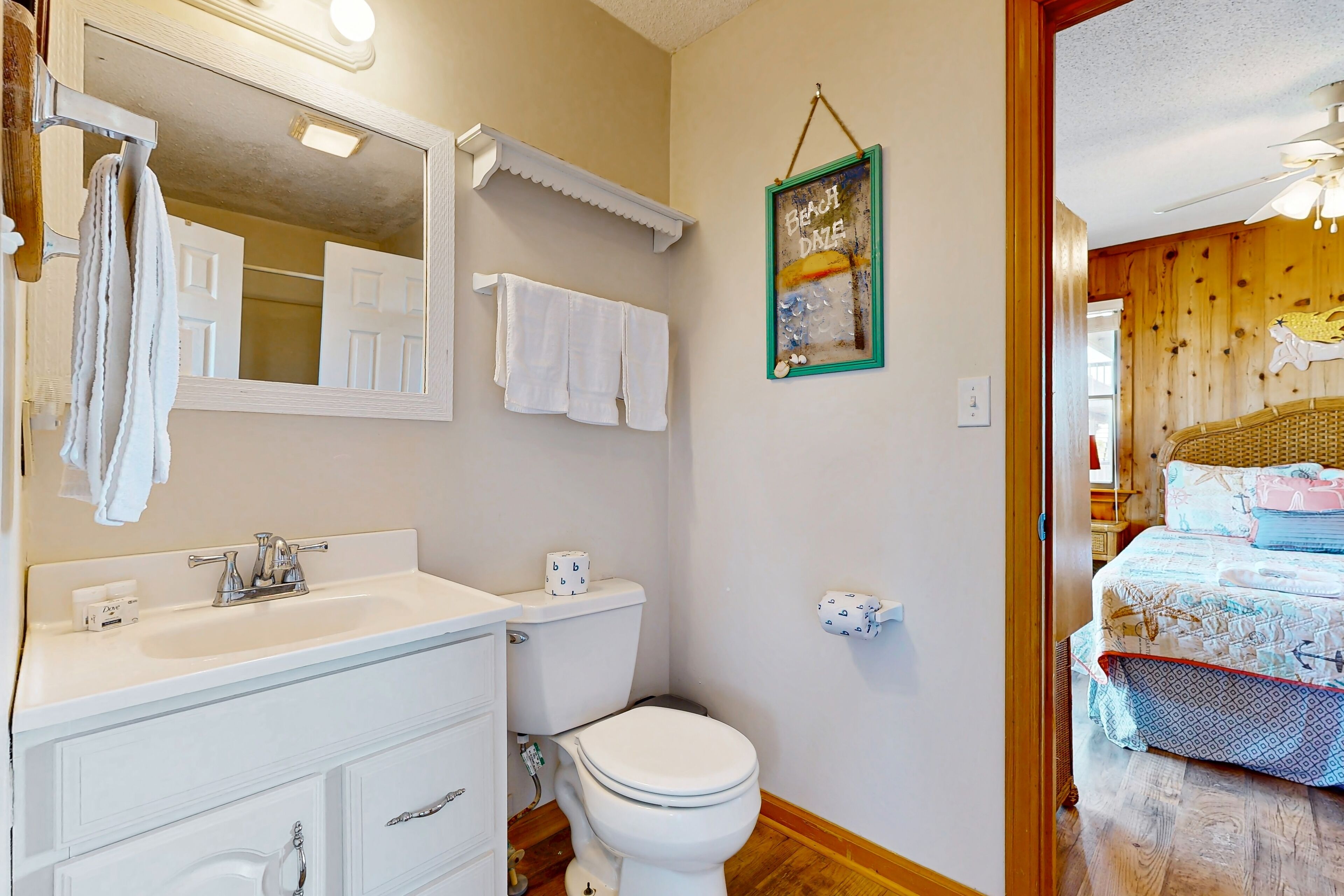 A bathroom with a white sink, mirror, and toilet. There is a towel rack with towels, a framed picture on the wall, and a wooden floor.