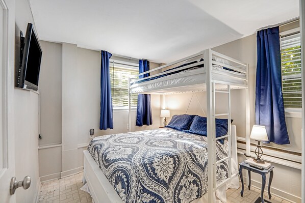 Un dormitorio con una cama individual superior e inferior, una colcha estampada, una mesita de noche con lámpara y un televisor instalado en la pared.