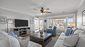 A modern living room with a flat-screen TV, grey sofas, a coffee table, and a ceiling fan. - Canal front home with a floating dock and large deck with sound views (Topsail Beach)