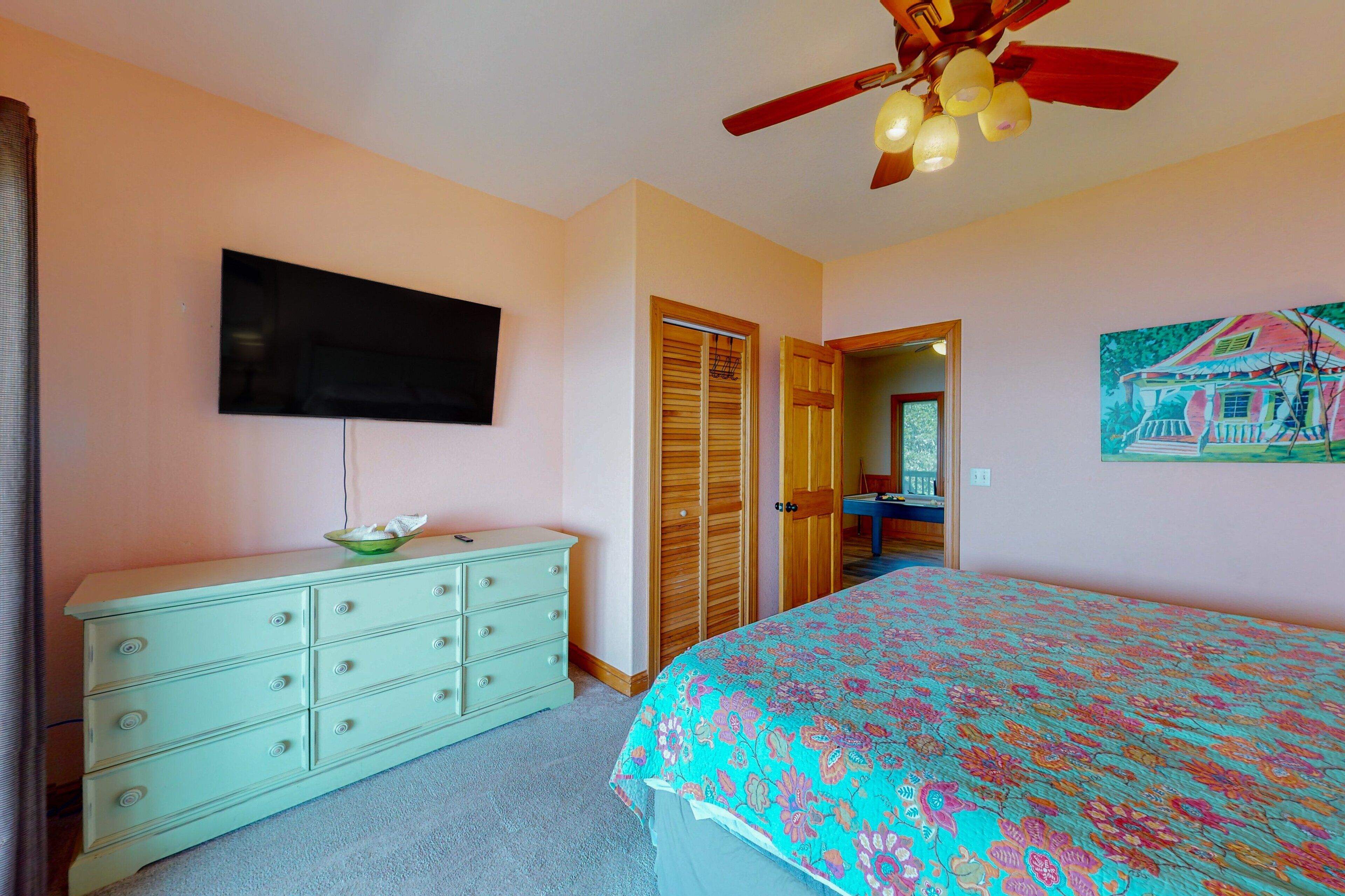A bedroom with a bed, a dresser, a television, a ceiling fan, and a door leading to another room.