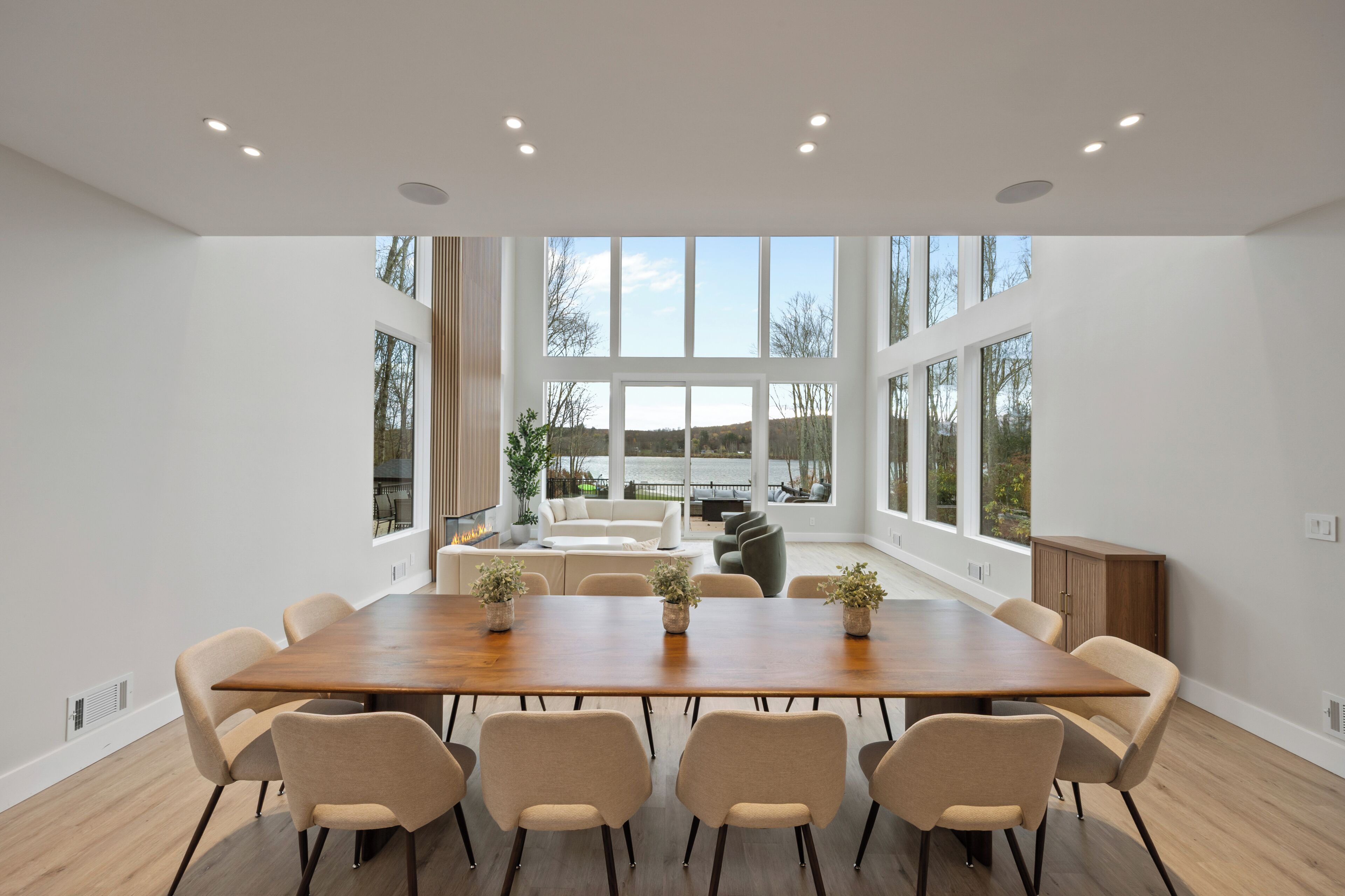 Un vaste espace dédié à la salle à manger, avec une longue table en bois, des chaises de couleur beige et de grandes fenêtres offrant une vue sur l’extérieur.