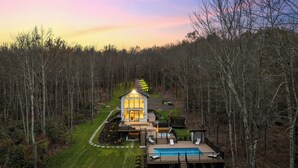 A modern house with a large deck and a swimming pool, surrounded by a forest.