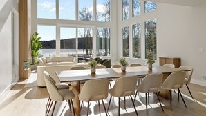 A spacious room with a large wooden table, six chairs, and a sofa. There are large windows offering a view of the outdoors.