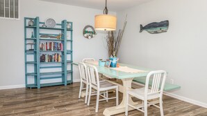 A dining area with a wooden table, white chairs, a teal bookshelf, and a wall clock. - Lovely beachfront condo with beach access, central AC, washer/dryer & fast WiFi (Carolina Beach)