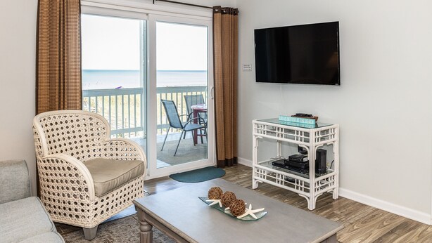 A living room with a wicker chair, a small table with decorative items, a white TV stand, and a view of the ocean through a sliding door. - Lovely beachfront condo with beach access, central AC, washer/dryer & fast WiFi (Carolina Beach)