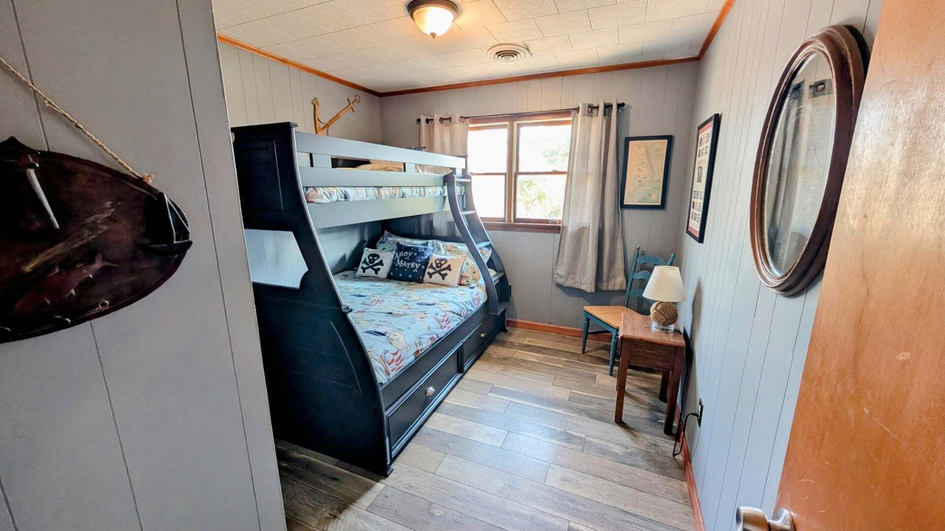 A room with a bunk bed, a small table with a lamp, a chair, and a window with curtains.