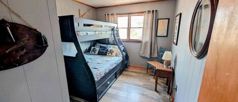 A room with a bunk bed, a small table with a lamp, a chair, and a window with curtains.