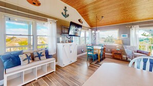 A spacious room with a wooden ceiling, large windows, a dining area, and a TV mounted on the wall. - Sea Escape 25: Beachfront, dog-friendly, private hot tub (Avon)