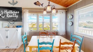 A dining area with a wooden table and chairs, a chandelier, and a view through large windows. - Sea Escape 25: Beachfront, dog-friendly, private hot tub (Avon)