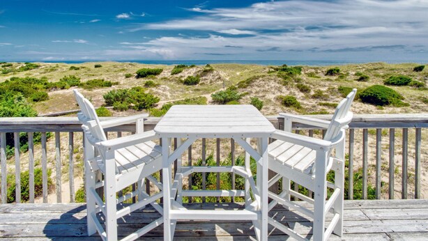 A wooden deck with a white table and two chairs, overlooking a beach with dunes and a clear sky. - Sea Escape 25: Beachfront, dog-friendly, private hot tub (Avon)