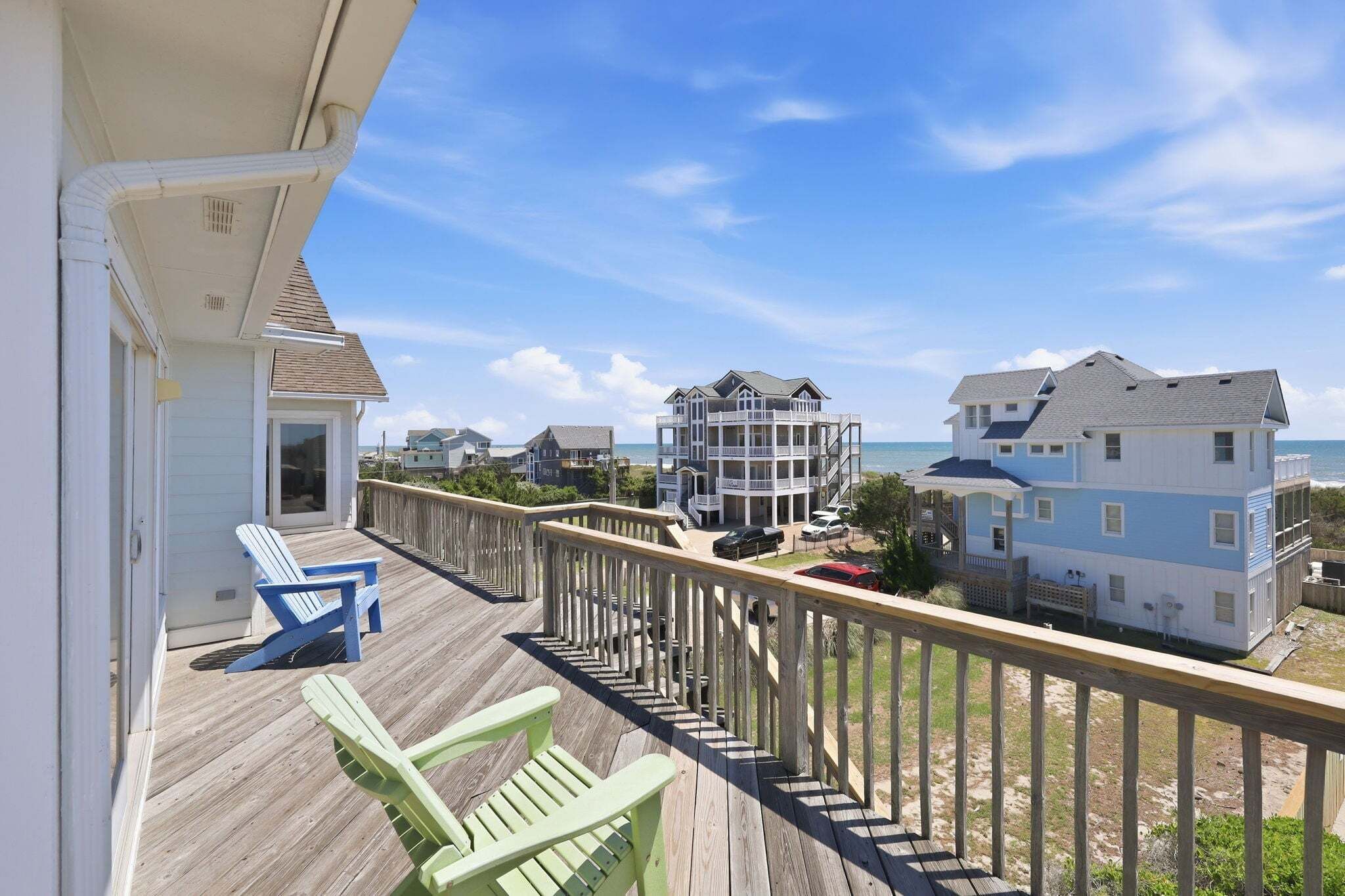 Un balcon avec deux fauteuils Adirondack, une terrasse en bois et une vue sur l’océan et les maisons voisines.