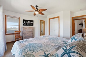 A bedroom with a bed, a ceiling fan, a dresser, a chair, a window with blinds, and a sign on the wall. - Ocean-view home with pool, hot tub, grill, decks & pool table (Waves)