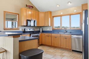 A kitchen with wooden cabinets, a stainless steel dishwasher, and a large window. - Ocean-view home with pool, hot tub, grill, decks & pool table (Waves)