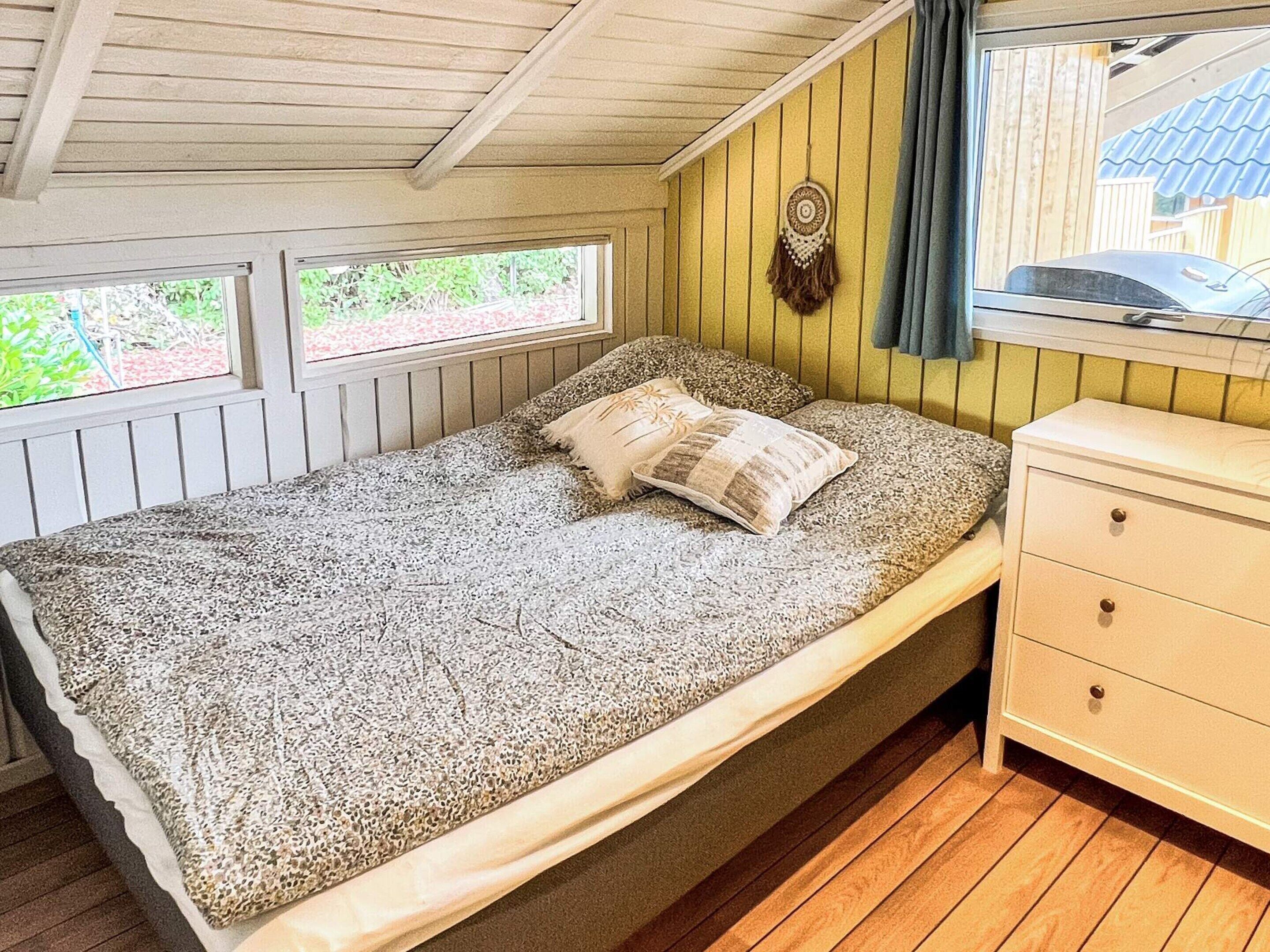 A single bed with a patterned bedspread, a bedside table with drawers, and a window with a view of greenery.