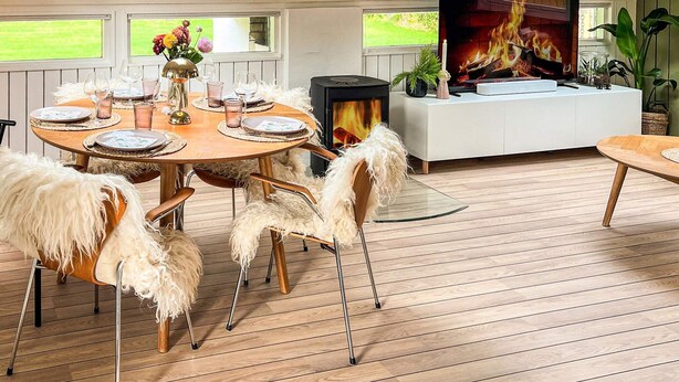 Ein gemütliches Zimmer mit einem runden Holzesstisch für vier Personen, einem weißen Sofa und einem Kamin, auf dessen Fernseher eine Feuerszene läuft.