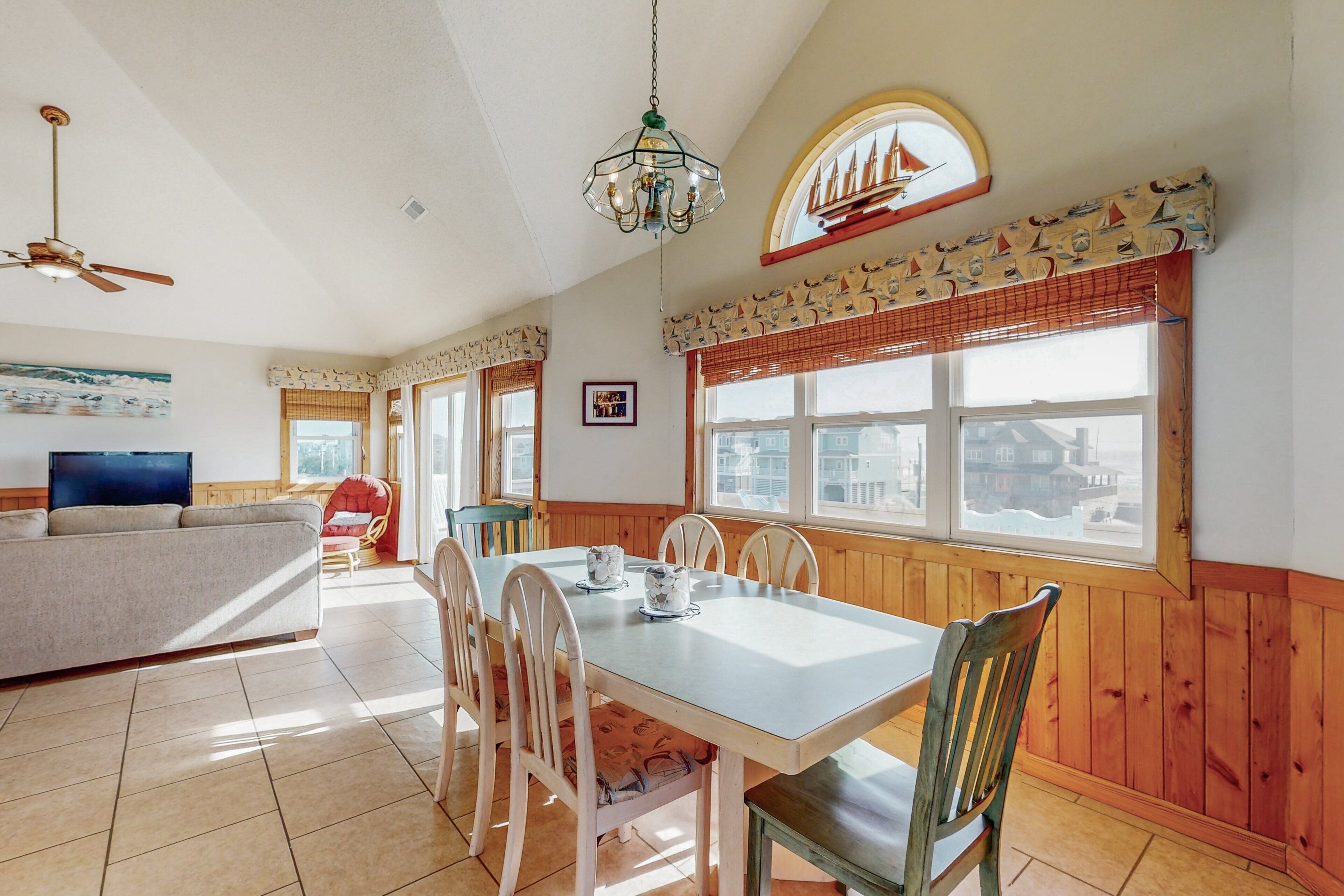 A dining area with a wooden table and chairs, a television, and a sofa in a room with wooden walls and large windows.