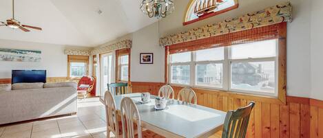 A dining area with a wooden table and chairs, a television, and a sofa in a room with wooden walls and large windows.