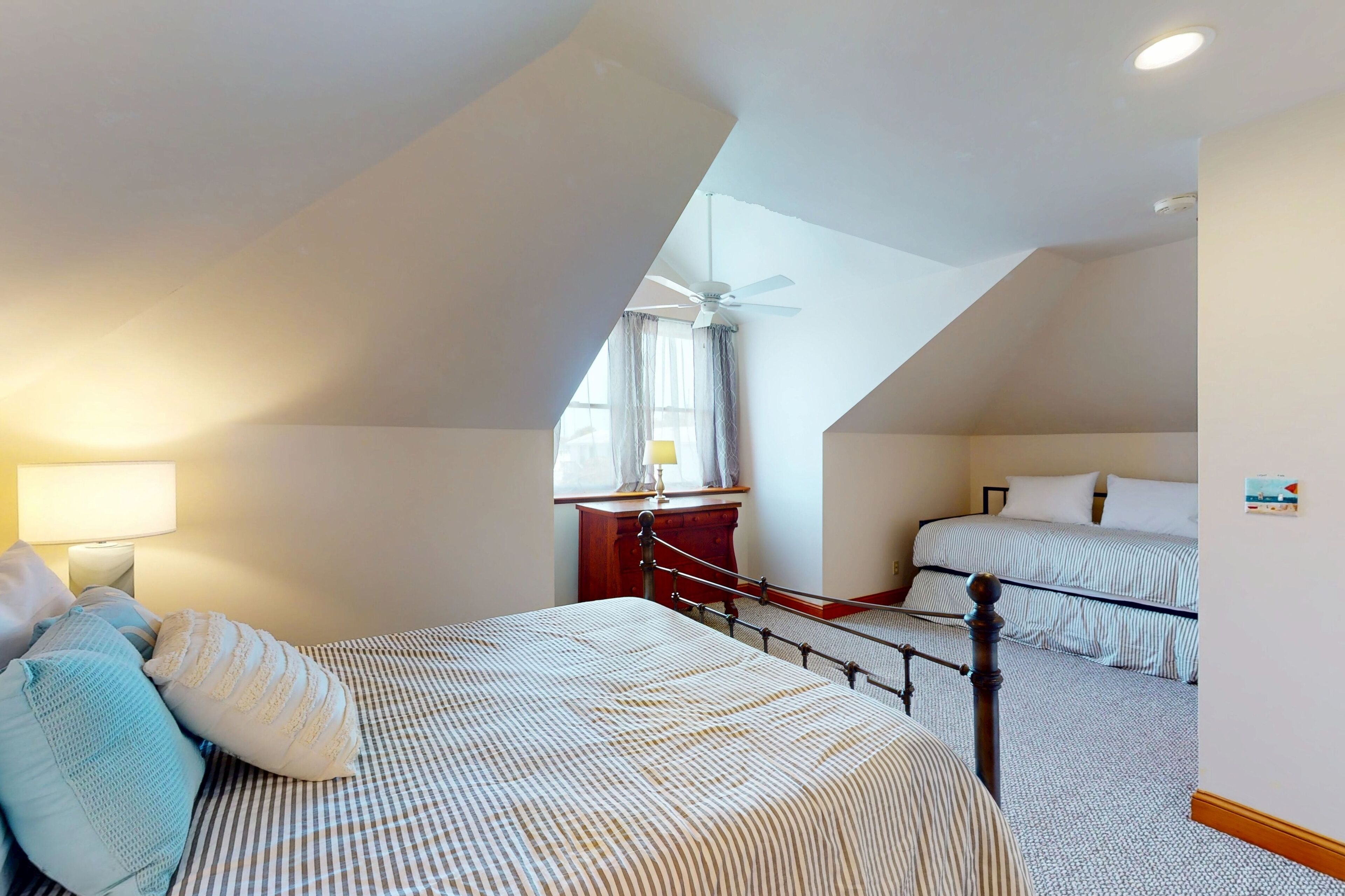 Un dormitorio con techo inclinado, una cama con ropa de cama a rayas, una mesita de noche con una lámpara y una ventana con cortinas.