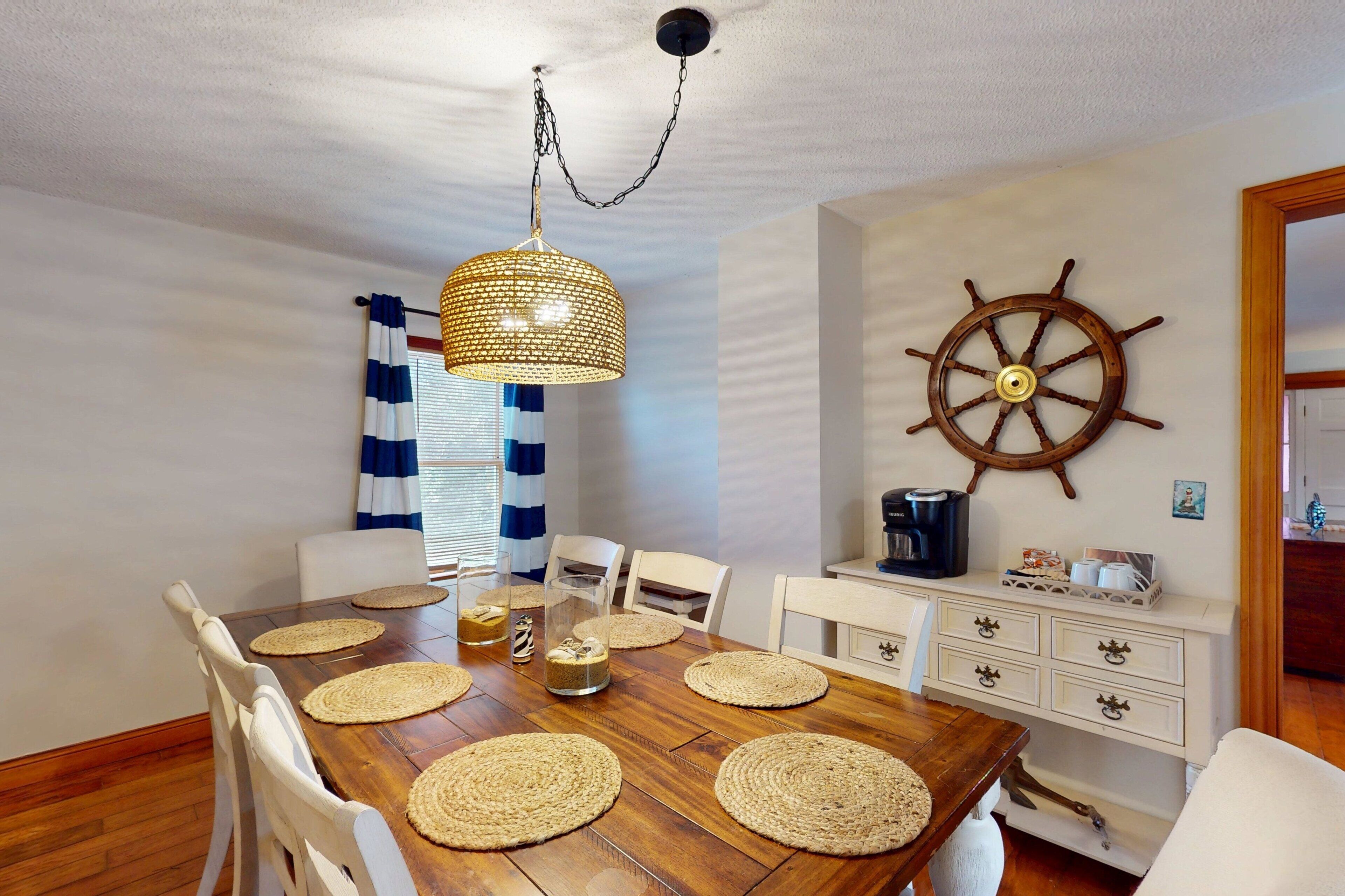 A dining area with a wooden table, white chairs, a ship's wheel decoration, and a hanging light fixture.