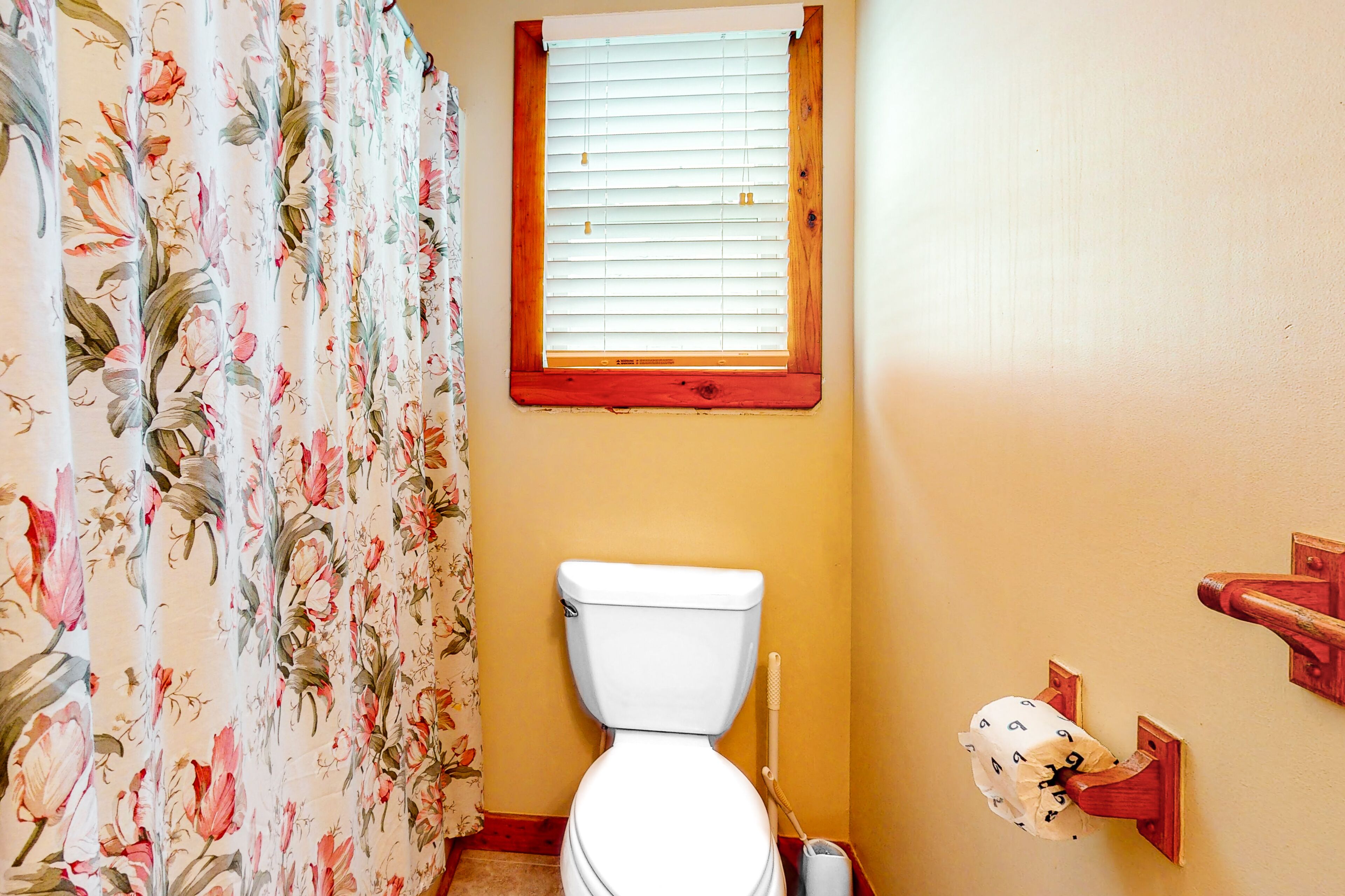 A bathroom with a toilet, a window with blinds, a shower curtain with floral patterns, and a toilet paper holder.