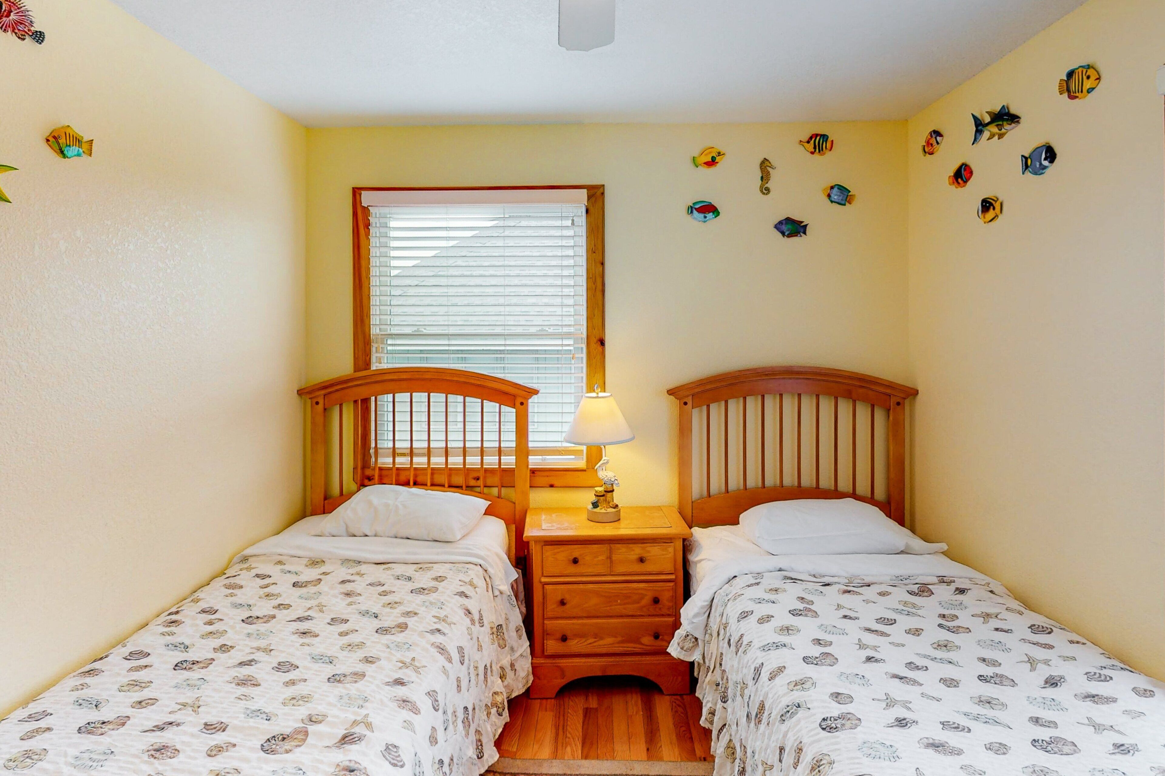 A room with two wooden beds, a window with blinds, a nightstand with a lamp, and wall decorations of fish and shells.