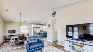 A spacious living room with a blue sofa, a dining area with a table and chairs, a kitchen in the background, and a large TV on a wooden stand. - Dog friendly coastal retreat with short walk to the beach and enclosed yard (Carolina Beach)