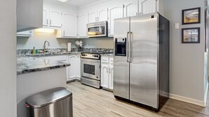 A modern kitchen with white cabinets, stainless steel appliances, a marble countertop, and a trash bin.