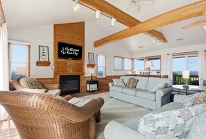 A spacious living room with a fireplace, wicker furniture, and a large window overlooking the sea. - Waterfront House w/ Private Hot Tub, Washer/Dryer, Shared Pool, Central AC, WiFi (Frisco)