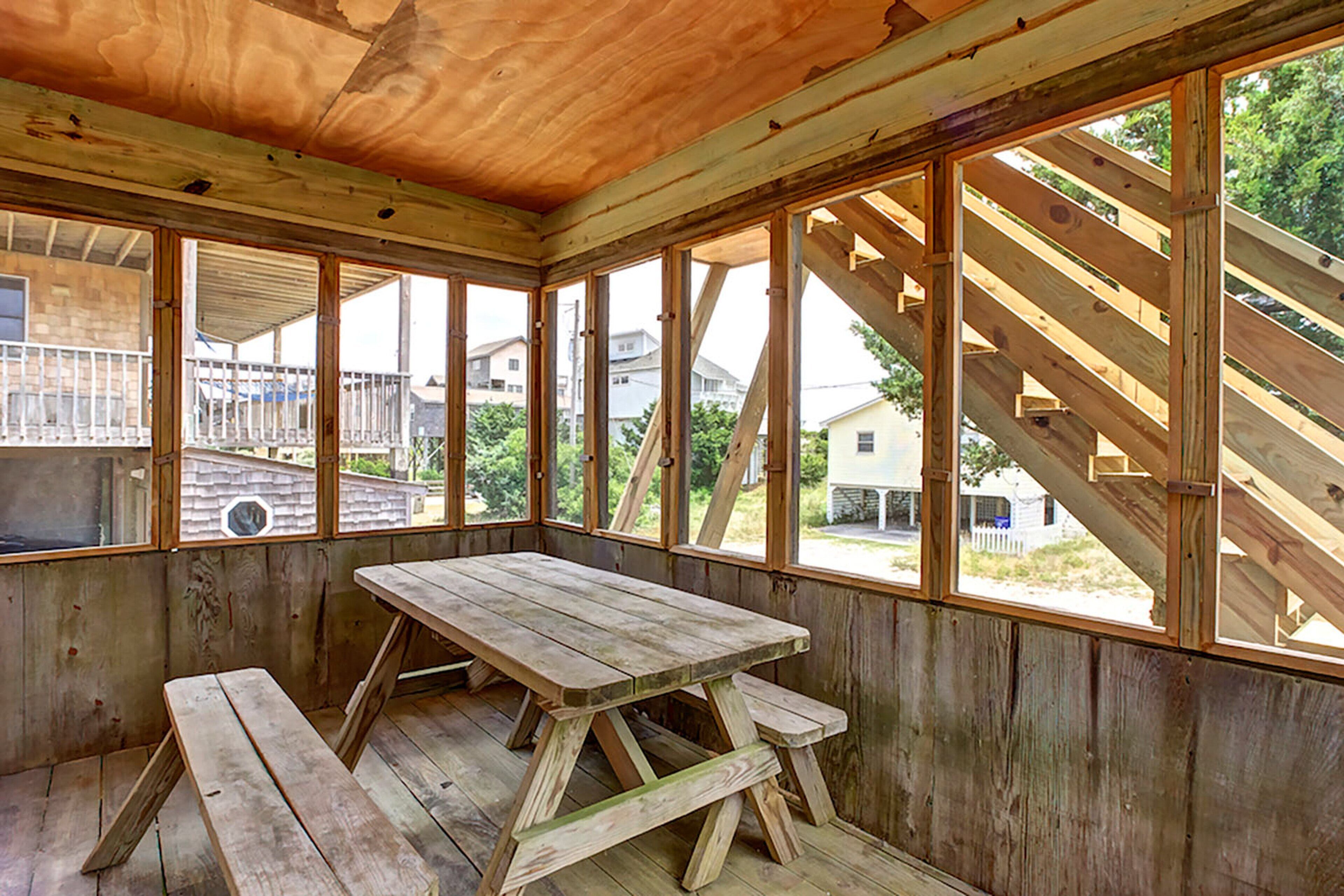 A wooden interior with a picnic table and benches, large windows, and a view of houses outside.