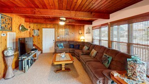 A cozy living room with wooden walls and ceiling, a sectional sofa, a coffee table, a television, and a window with blinds. - Beautiful house w/Shared Pool, Private Washer/Dryer, Central AC, Free WiFi (Buxton)